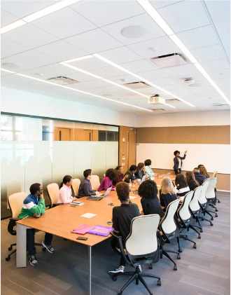 15 people of mixed color and age sitting around a large conference table. One person is presenting something in the front of the room.